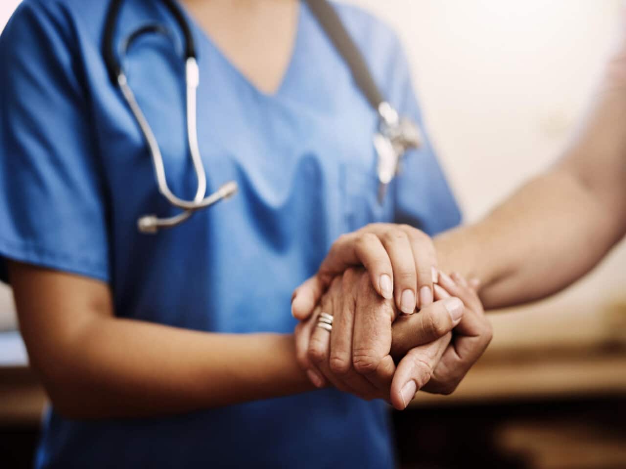 profissional formada em enfermagem do trabalho com um estetoscópio no pescoço, segurando a mão de uma paciente durante um atendimento.