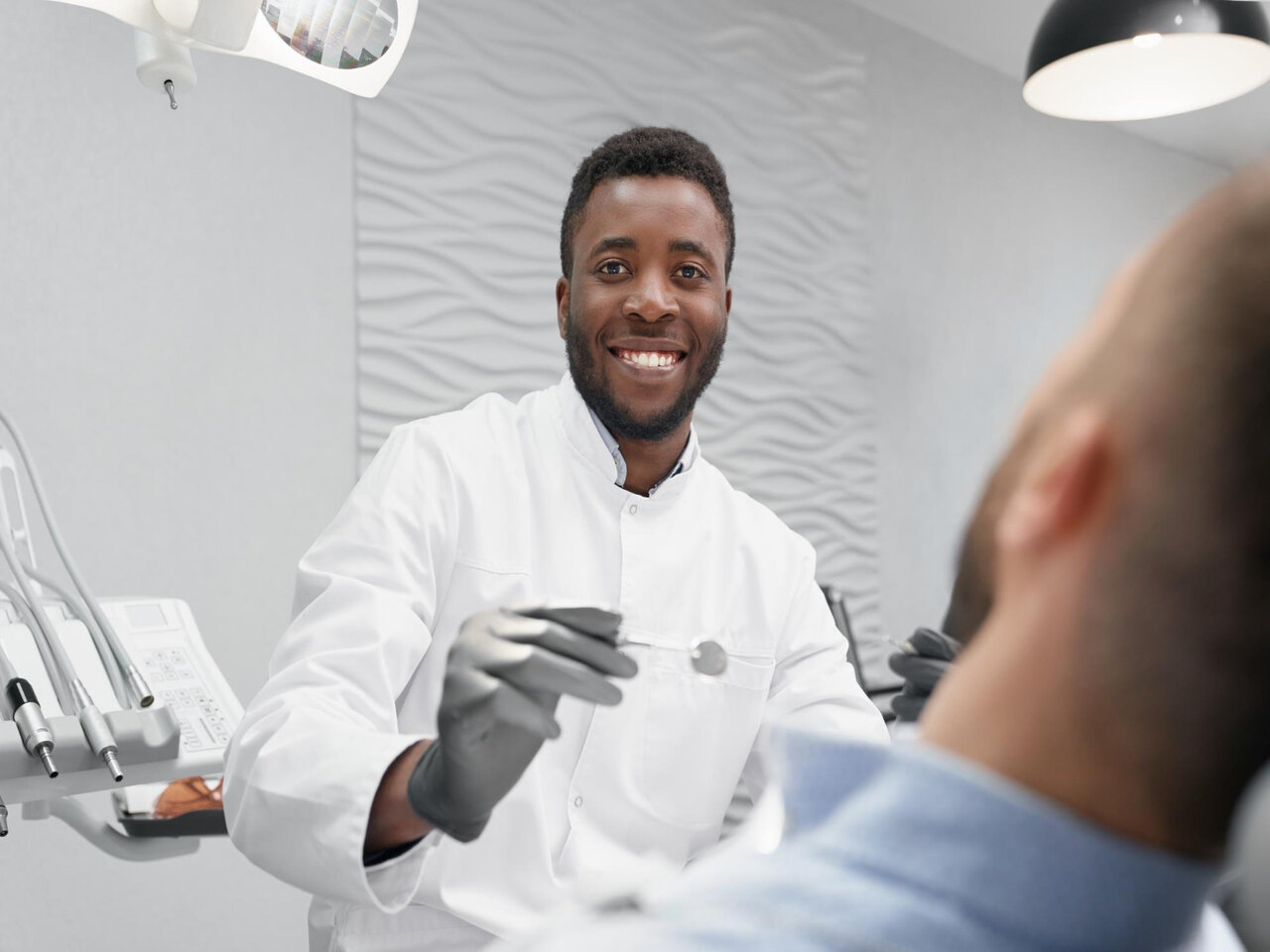 Faculdade de Odontologia: dentista em seu consultório, segurando uma pequena espátula enquanto atende um paciente deitado na maca.