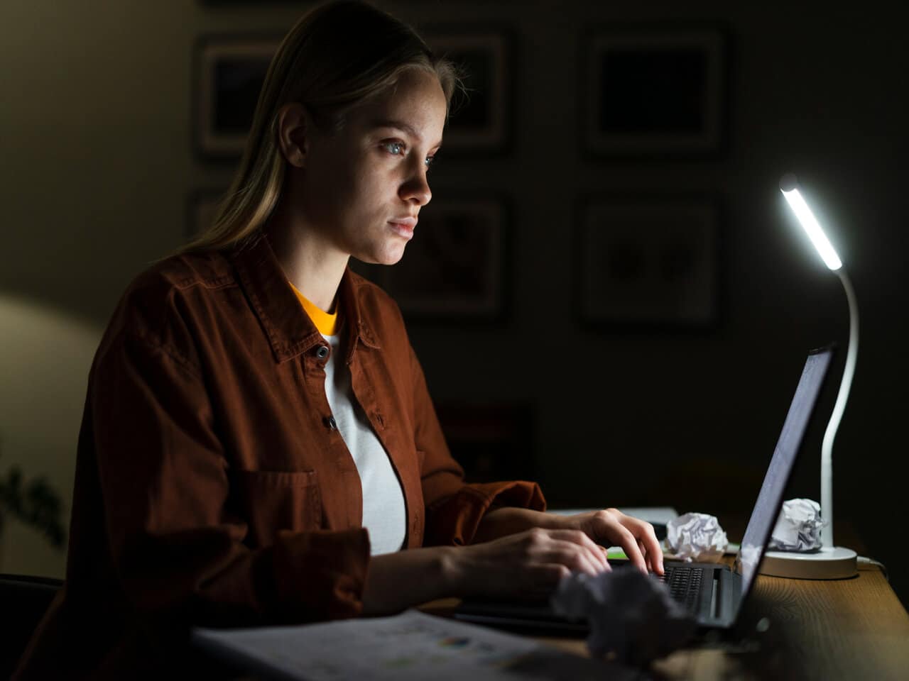 Mulher jovem em sua mesa de estudos iluminada por um abajur, utilizando um notebook para estudar à noite. Em cima da mesa, alguns papéis amassados e um bloco de folhas.