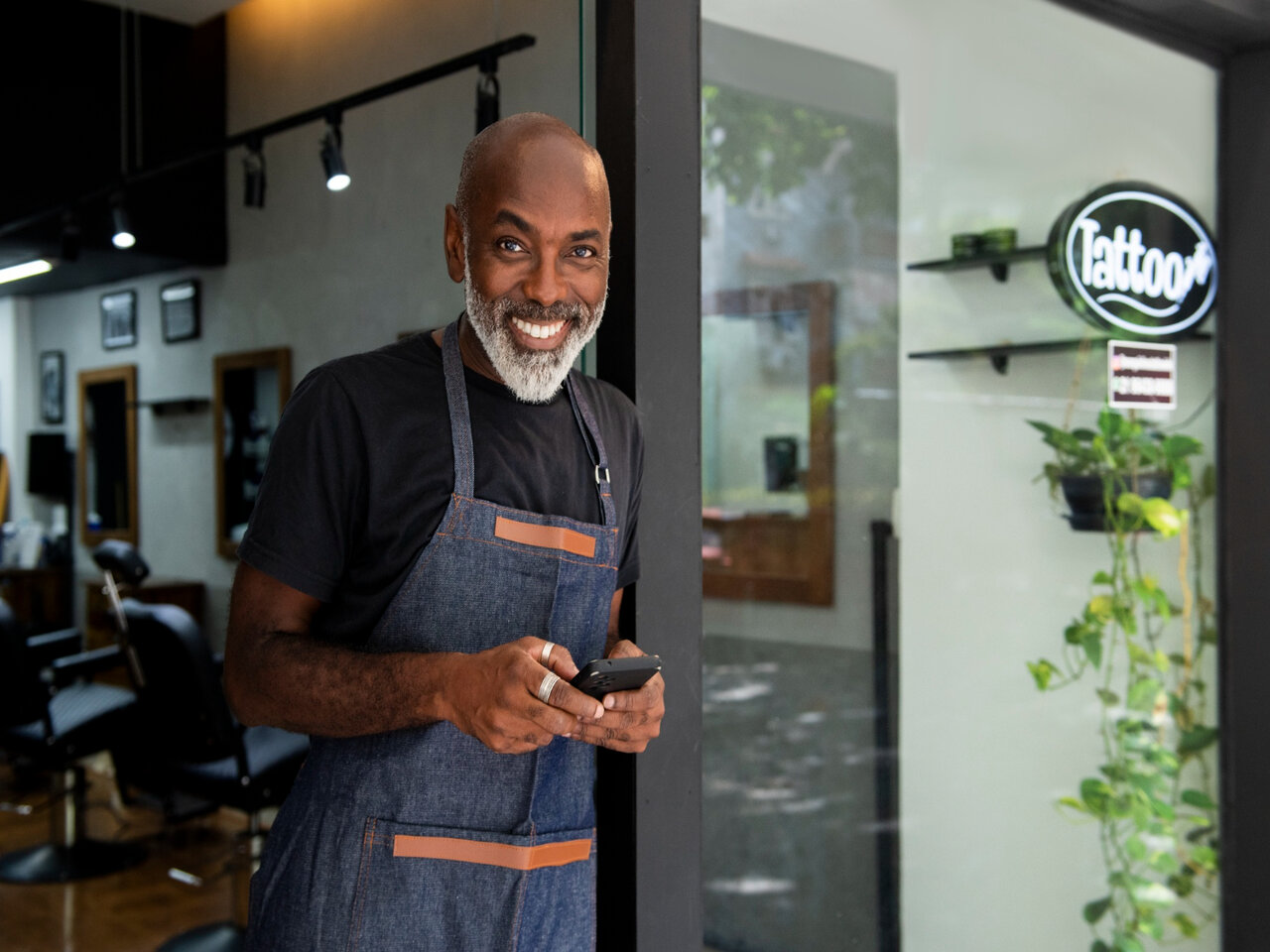 Atividades MEI: proprietário de um salão em seu ambiente de trabalho sorrindo para a câmera enquanto segura um celular.