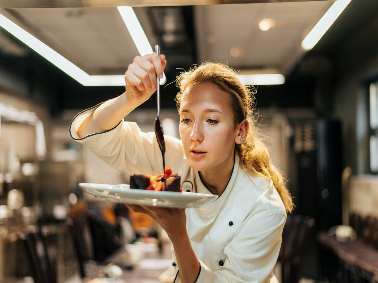Mulher chef de cozinha utilizando uma dolmã, segurando um prato com uma receita que contém carne e despejando um molho por cima por meio de uma colher.