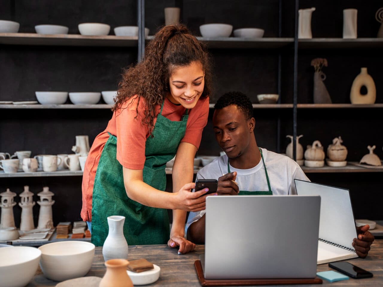 Vantagens de ser MEI: microempreendedor e sua funcionária em uma pequena loja de artesanato, consultando alguma informação no celular. Na mesa à sua frente, algumas peças de porcelana e um notebook aberto.