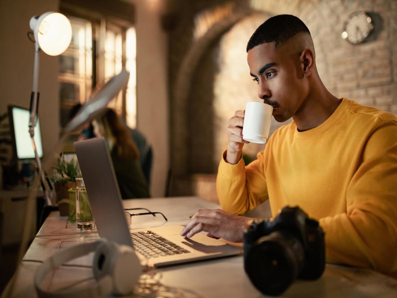 Microempreendedor individual em seu estúdio de fotografia, tomando uma xícara de café enquanto utiliza o computador para emitir o boleto MEI. Em cima de sua mesa estão uma câmera fotográfica, um fone de ouvido, um copo com água e um óculos.