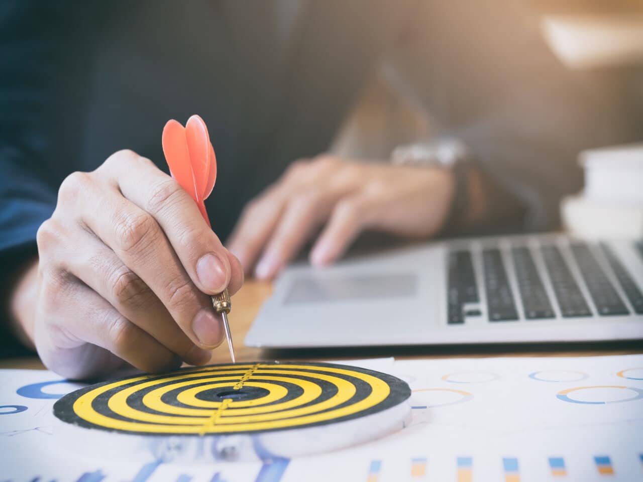 Dia do Profissional de Marketing: profissional de marketing em seu escritório segurando um dardo e apontando-o para o alvo em cima de sua mesa. Ao seu lado, um notebook usado para trabalhar.