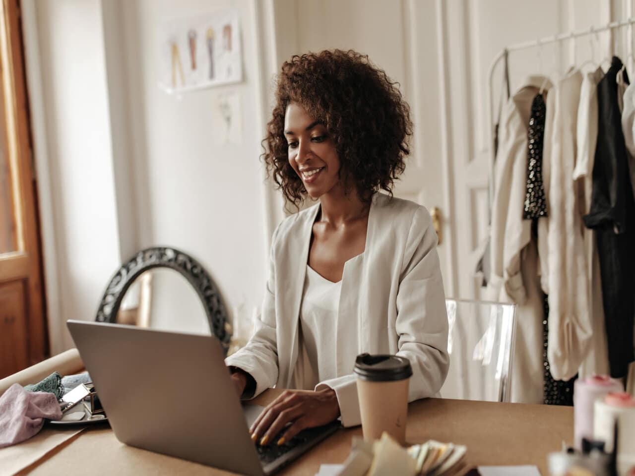 Empreendedorismo digital: empreendedora em seu ateliê, utilizando um notebook para trabalhar. Ao fundo, algumas roupas em uma arara e, sobre a mesa, um copo de café e pedaços de tecido.