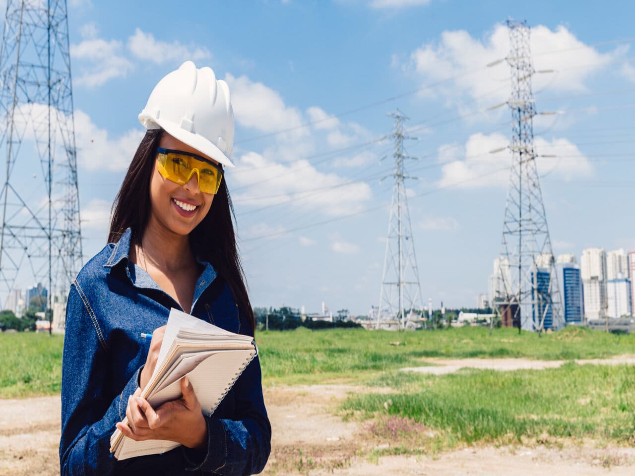 Estágio em engenharia elétrica: estagiária com um capacete branco, em uma área aberta com torres de alta voltagem, utilizando um lápis e um caderno para fazer anotações.