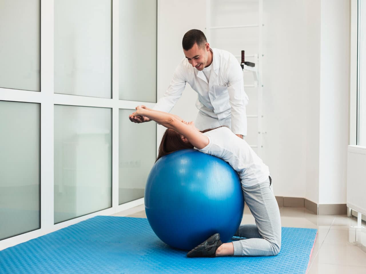 Áreas da fisioterapia: fisioterapeuta e paciente em um consultório durante uma sessão de fisioterapia. Ele está auxiliando a paciente a fazer o exercício em uma bola suíça.