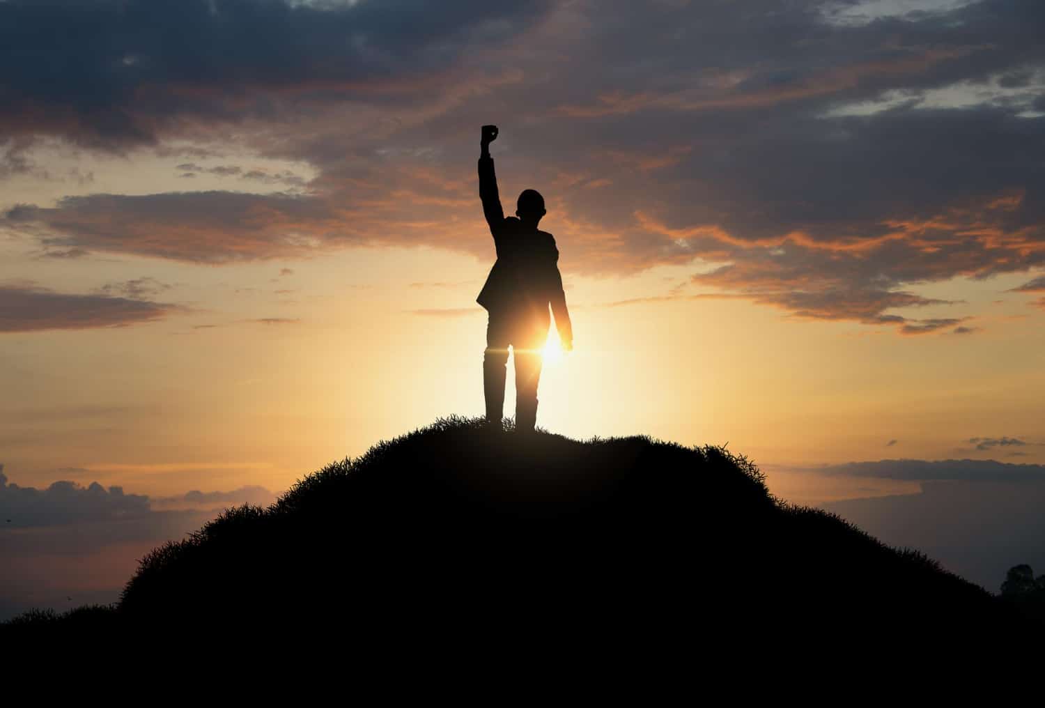 Silhueta de um homem com o braço esquerdo levantado, no topo de uma montanha, durante um pôr do sol. Ele conquistou o objetivo após desenvolver a autodisciplina.
