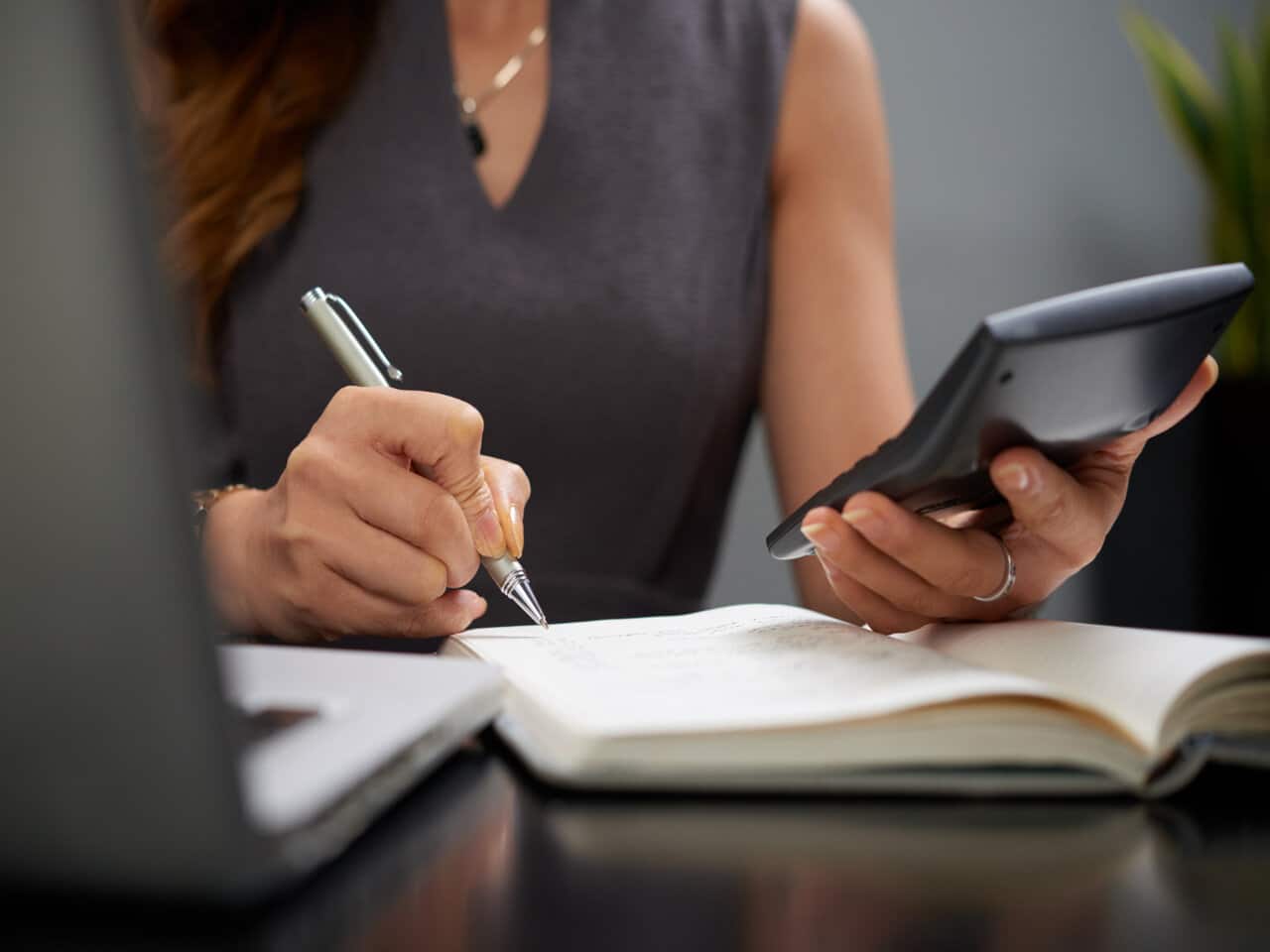 Dia do Profissional de Contabilidade: contadora em seu escritório utilizando uma calculadora para fazer contas, enquanto usa uma caneta para fazer anotações em um caderno.