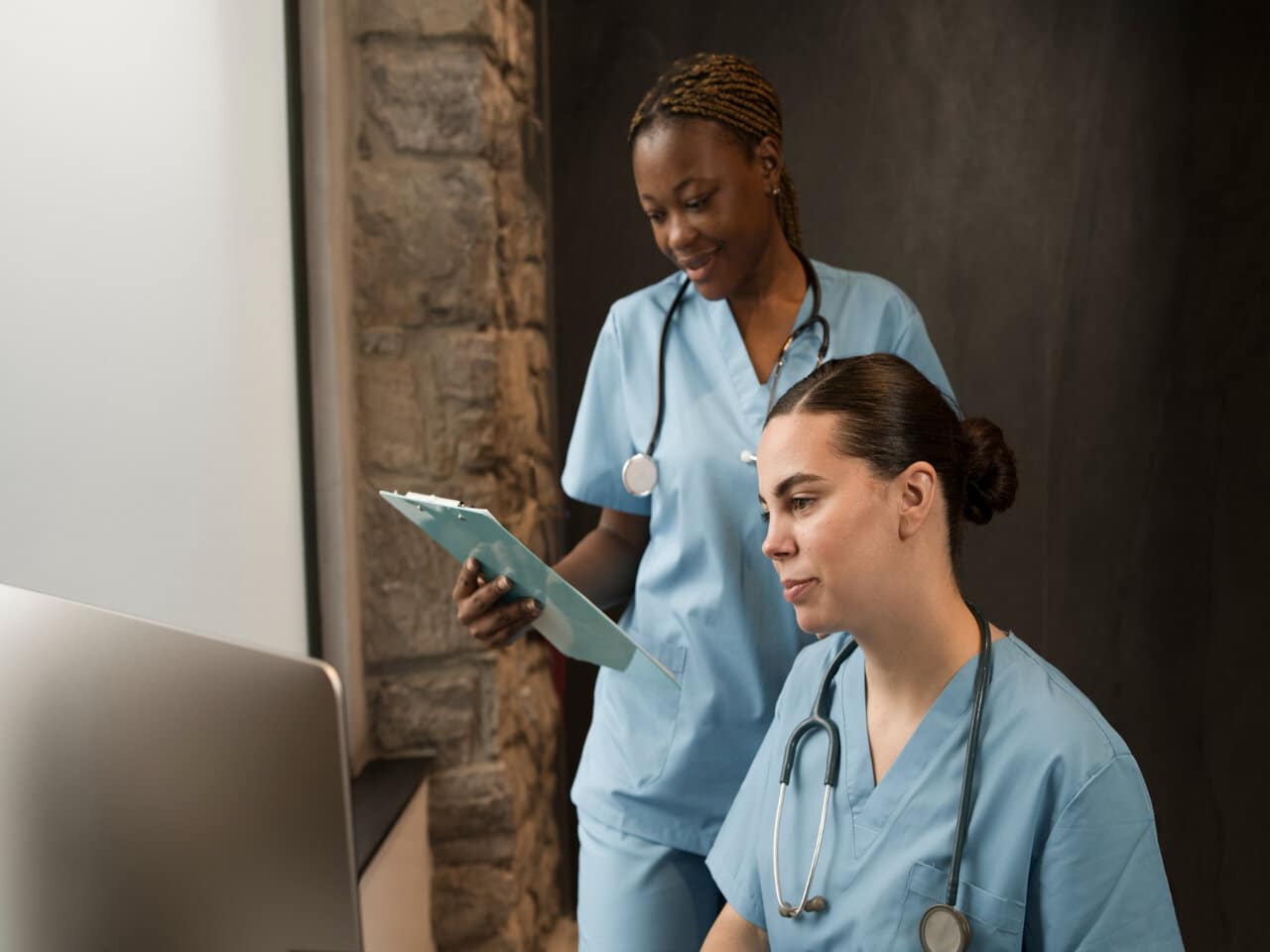 Estágio em enfermagem: duas enfermeiras em um consultório, utilizando jaleco azul e estetoscópio no pescoço. Uma está sentada de frente para um computador, enquanto a outra está em pé ao seu lado, segurando uma prancheta.