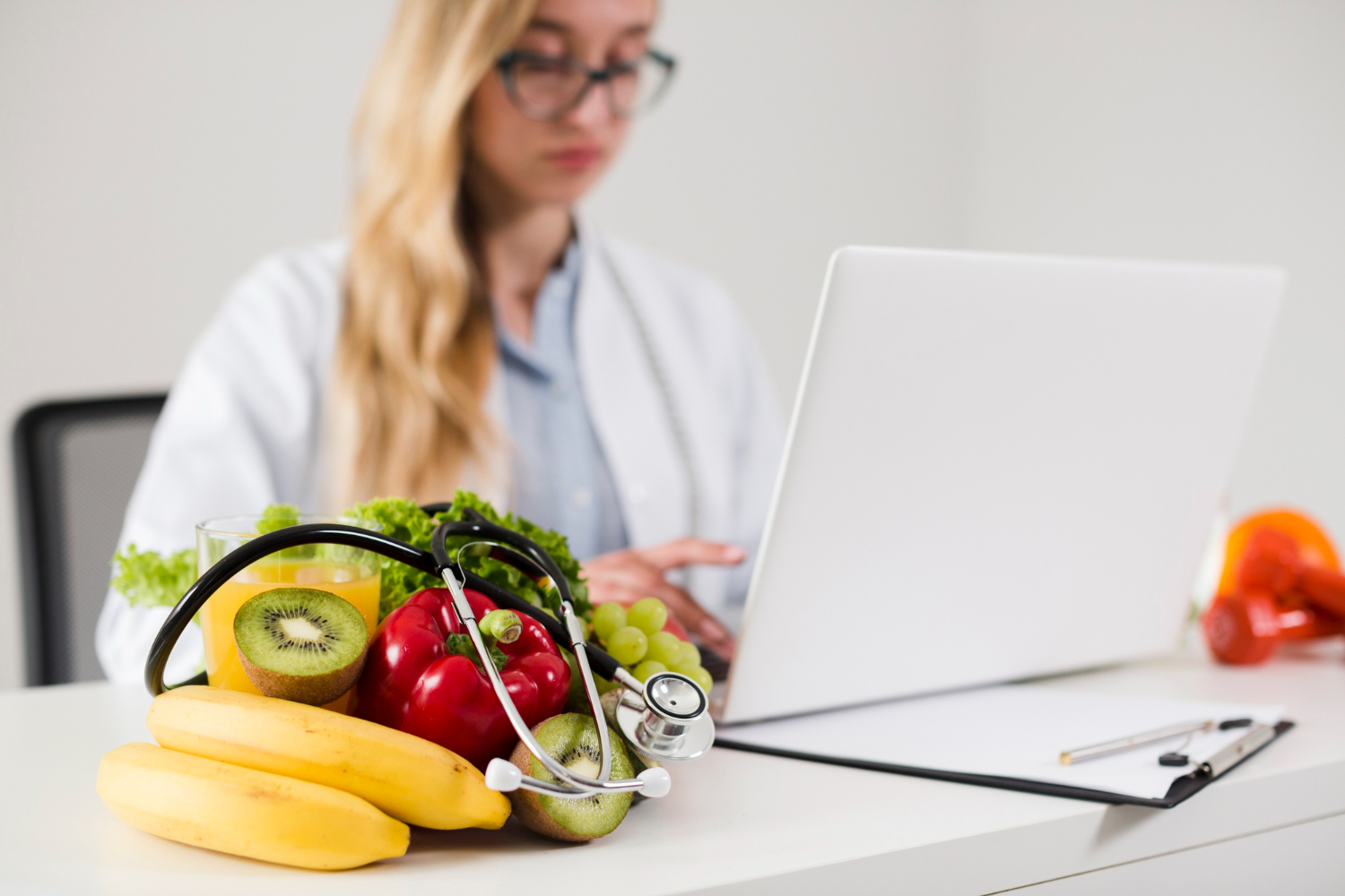 Estagiária em um consultório, utilizando um notebook durante o estágio em nutrição. Na mesa à sua frente, algumas frutas empilhadas com um estetoscópio em cima.