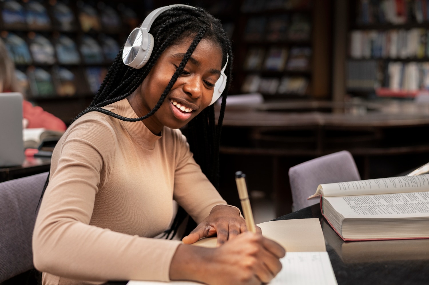 Jovem mulher estudante em uma biblioteca, com alguns livros abertos à sua volta, fazendo anotações em um caderno e ouvindo músicas para estudar por meio de um fone de ouvido.
