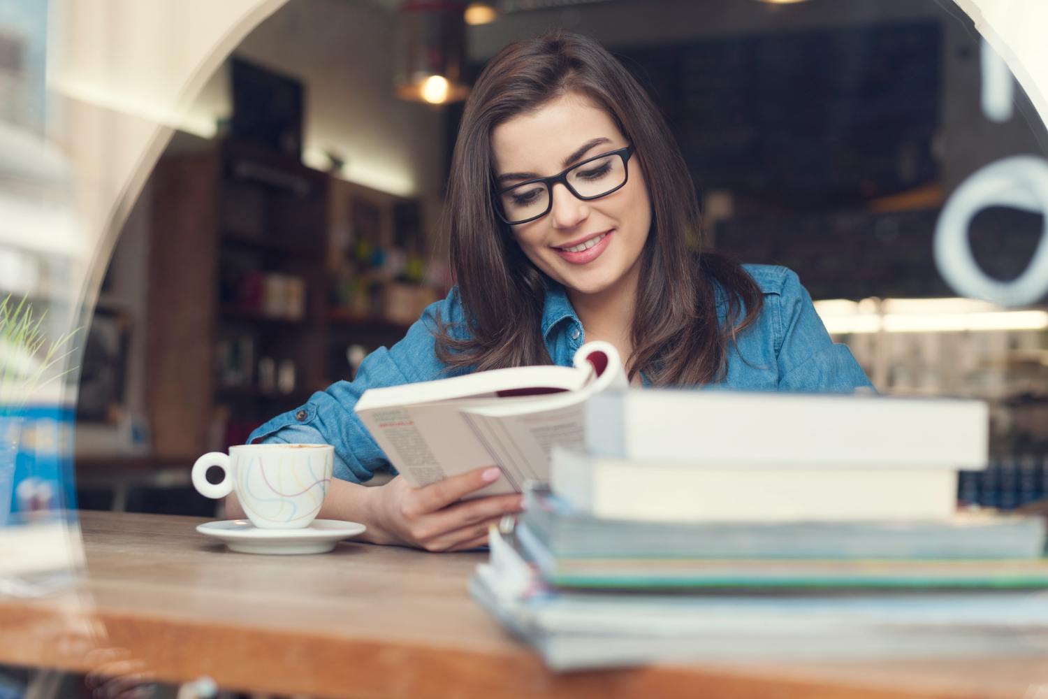 Aluna sentada em uma mesa de estudos dentro da biblioteca da faculdade, lendo um livros de terapia ocupacional. Ao seu lado, uma pilha de livros e uma xícara de café.