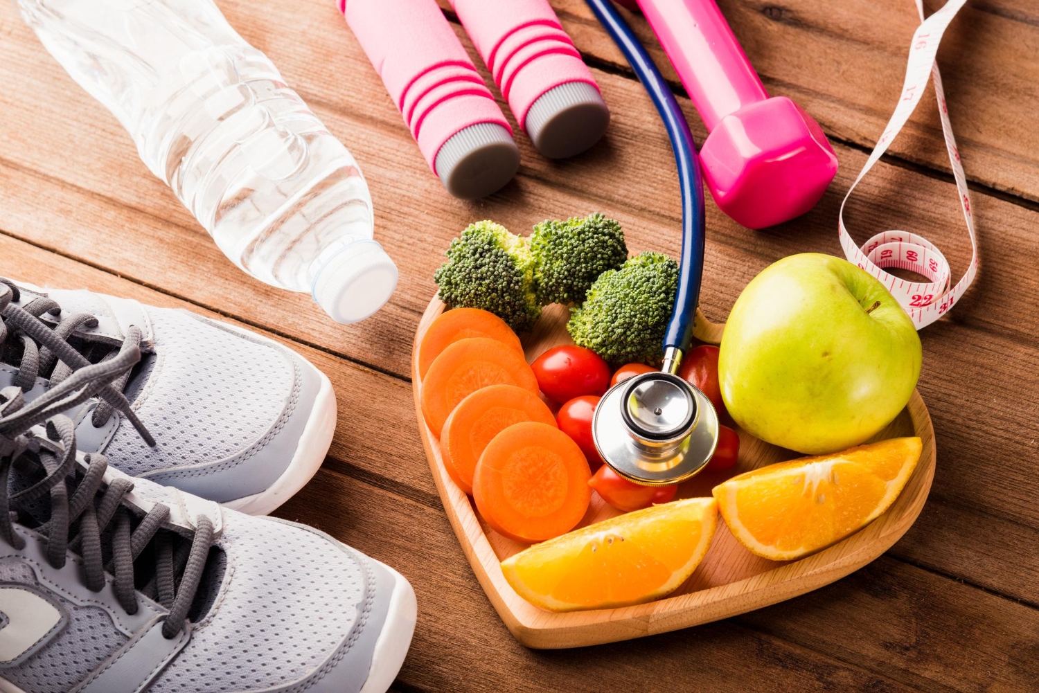 Cesta em formato de coração com frutas dentro, em cima de uma mesa, com um estetoscópio sobre elas. Ao lado, um par de tênis, halteres, uma garrafa de água e uma fita métrica, representando a nutrição esportiva.