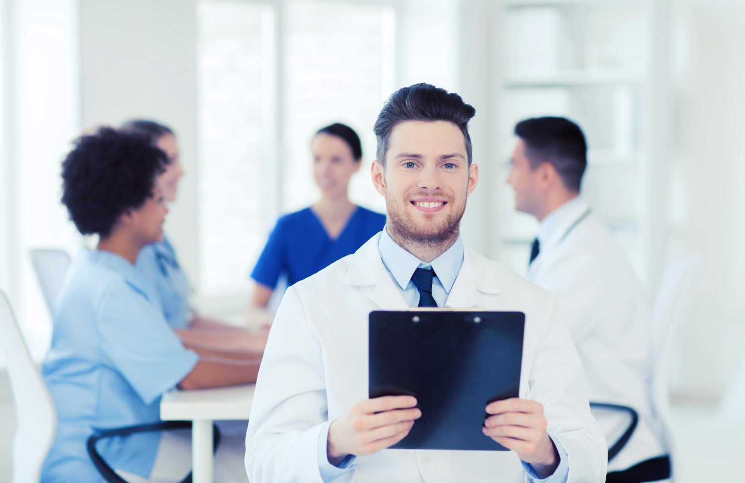 Técnico em Gerência em Saúde em um escritório segurando um tablet. Ao fundo, alguns funcionários do hospital sentados em suas cadeiras, durante uma reunião.