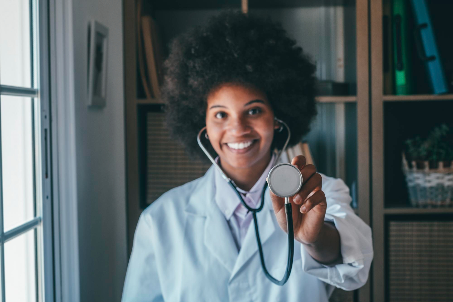 O que é ser médico: médica em seu consultório utilizando um estetoscópio, enquanto sorri para a câmera e exibe parte do equipamento