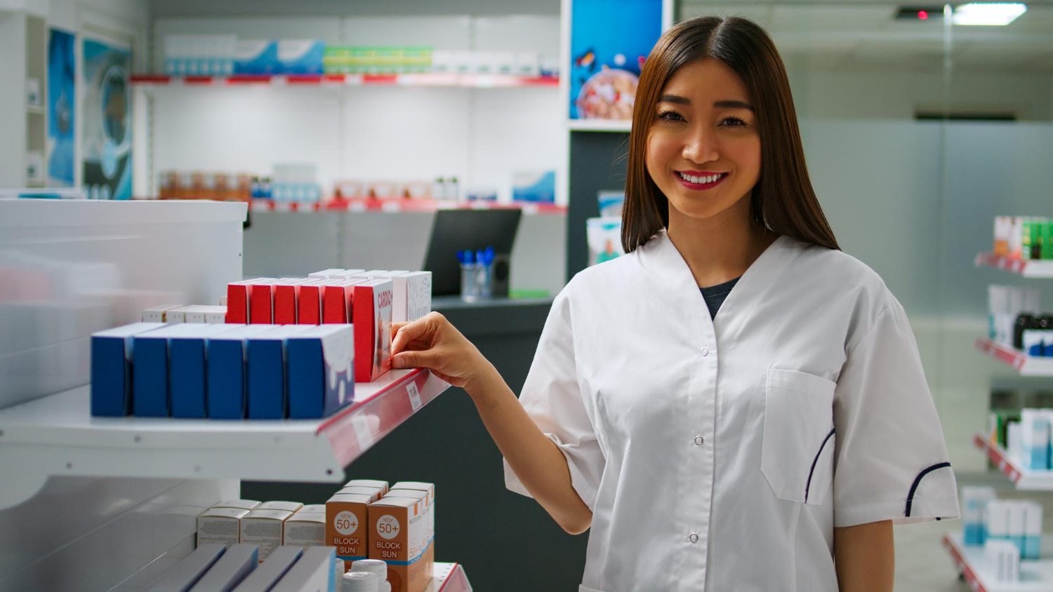 Grade curricular de farmácia: estudante de farmácia em um laboratório farmacêutico, olhando sorridente para a câmera enquanto segura uma das caixas de medicamentos na prateleira ao lado dela.