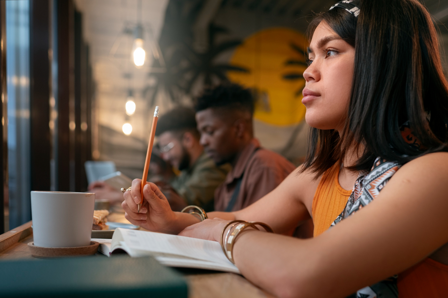 Jovem estudante em uma mesa de estudos, segurando um lápis e com um caderno aberto à sua frente. Ela está com uma expressão pensativa, enquanto escolher entre a área de exatas ou humanas.