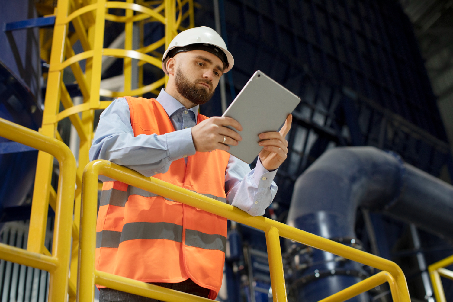 Engenheiro em uma fábrica utilizando capacete branco e colete laranja, enquanto coloca em prática seu inglês técnico ao ler um manual de instruções em um tablet.