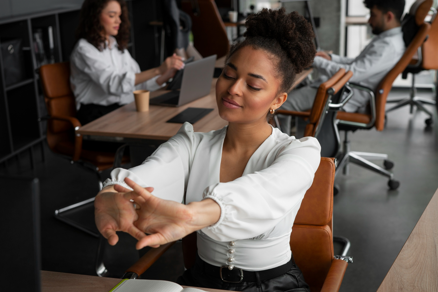Mulher em um escritório, sentada em sua cadeira enquanto estica as mãos para a frente em um movimento de alongamento dos dedos. Ela está fazendo uma pausa para o descanso após aumentar sua produtividade no trabalho.