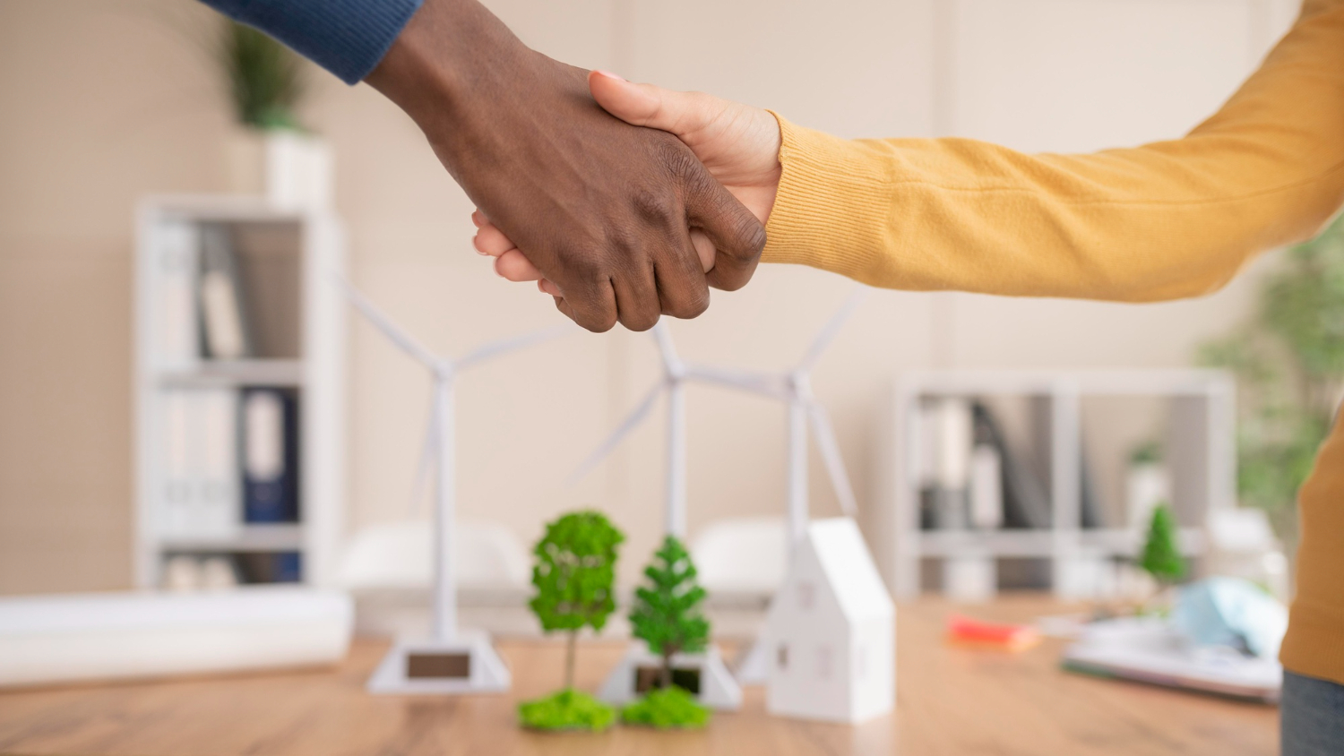 Imagem de um aperto de mãos entre duas pessoas, selando um acordo de comercialização de energia elétrica. Ao fundo, em cima de uma mesa, algumas maquetes de turbinas eólicas e pequenas árvores.