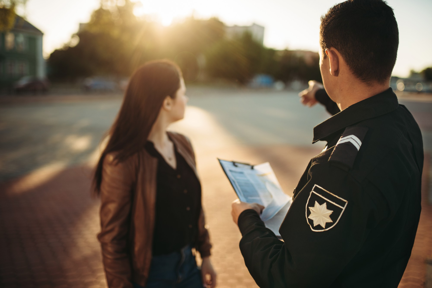 Agente de segurança pública segurando uma prancheta, enquanto aponta para um estacionamento, dando orientação a uma mulher.