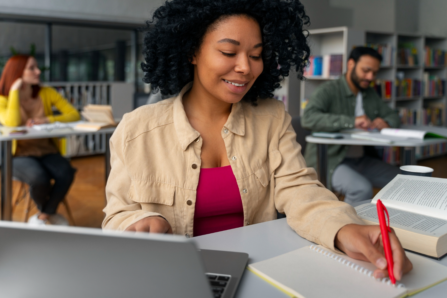 Estudante em uma biblioteca de faculdade, utilizando um notebook e um livro para estudar a grade curricular de Administração