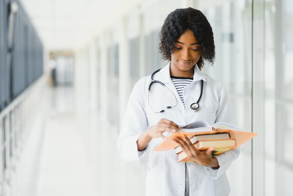 Jovem estudante da área da saúde, em um corredor de uma instituição, com um estetoscópio pendurado no pescoço enquanto segura alguns livros de medicina.