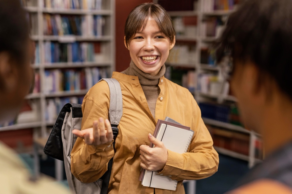 Orientador de TCC: professora em uma biblioteca de faculdade, com uma mochila nas costas e segurando um caderno enquanto gesticula e conversa com dois alunos que buscam orientação.