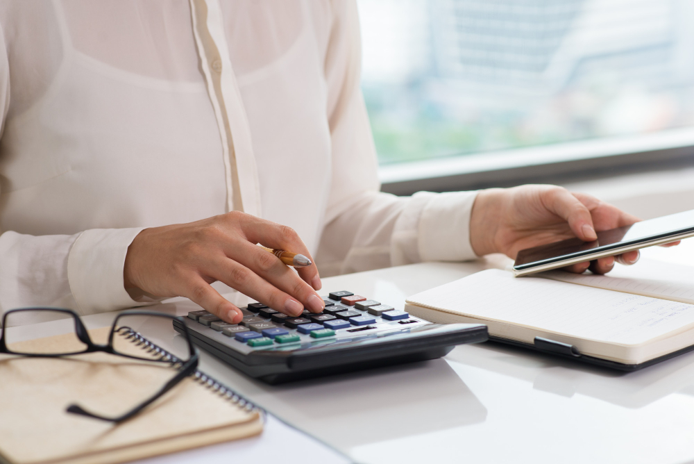 Áreas de exatas: gestora de finanças em um escritório, sentada em sua mesa de trabalho utilizando uma calculadora e um celular. À sua frente, dois cadernos para anotações e um óculos.