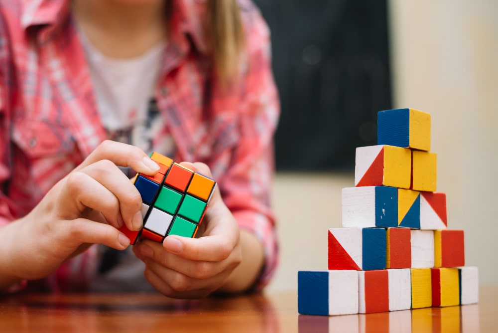 Jovem menina em uma sala utilizando o raciocínio lógico para resolver um cubo mágico. Na mesa à sua frente, uma pirâmide feita com pequenos blocos de madeira coloridos.