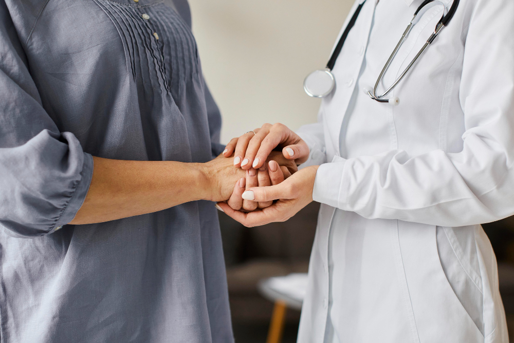 Médica com especialização multiprofissional em oncologia com um estetoscópio no pescoço, segurando as mãos de uma paciente em sina de solidariedade
