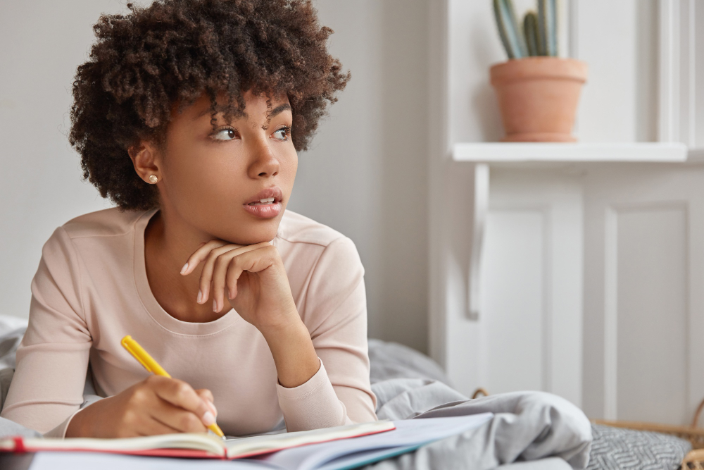 Jovem estudante com a mão no queixo, pensativa, buscando maneiras de aplicar a coerência textual no texto em que está escrevendo em seu caderno de estudos