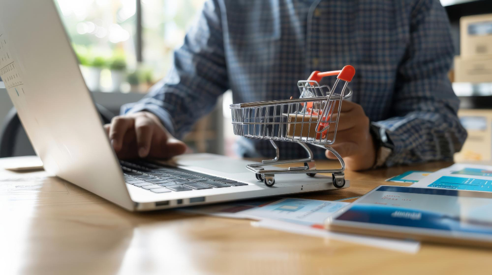 Dono de uma loja virtual formado na faculdade de E-commerce trabalhando em um notebook. Ao seu lado, uma miniatura de um carrinho de supermercado, representando as vendas online.