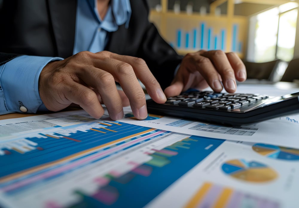 Profissional formado na faculdade de Gestão Financeira em seu escritório utilizando uma calculadora para fazer cálculos. Em cima da mesa, alguns documentos e relatórios com gráficos.