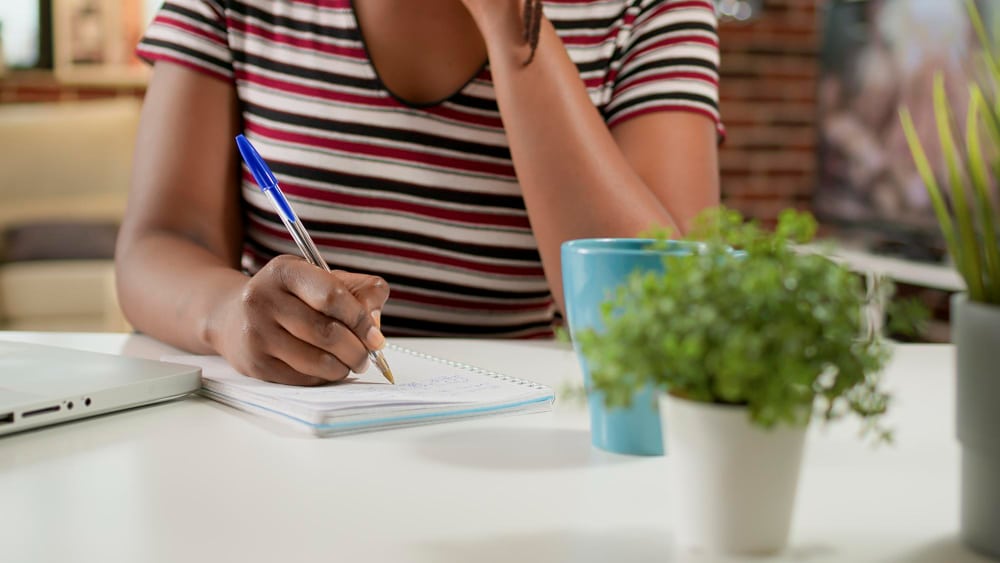 O que é argumento: jovem estudante utilizando uma caneta para escrever em um caderno em uma mesa, enquanto treina para a redação do ENEM
