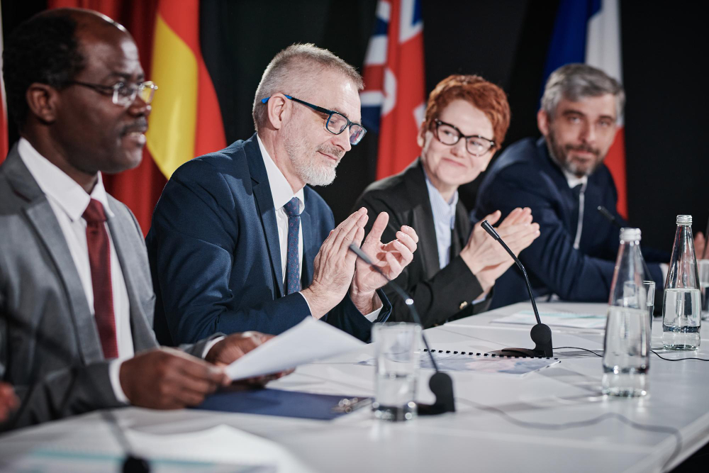 Integrantes de um dos blocos econômicos internacionais reunidos em uma mesa, ouvindo o discurso do representante de um dos países. Ao fundo, algumas bandeiras que representa as nações que fazem parte do bloco.