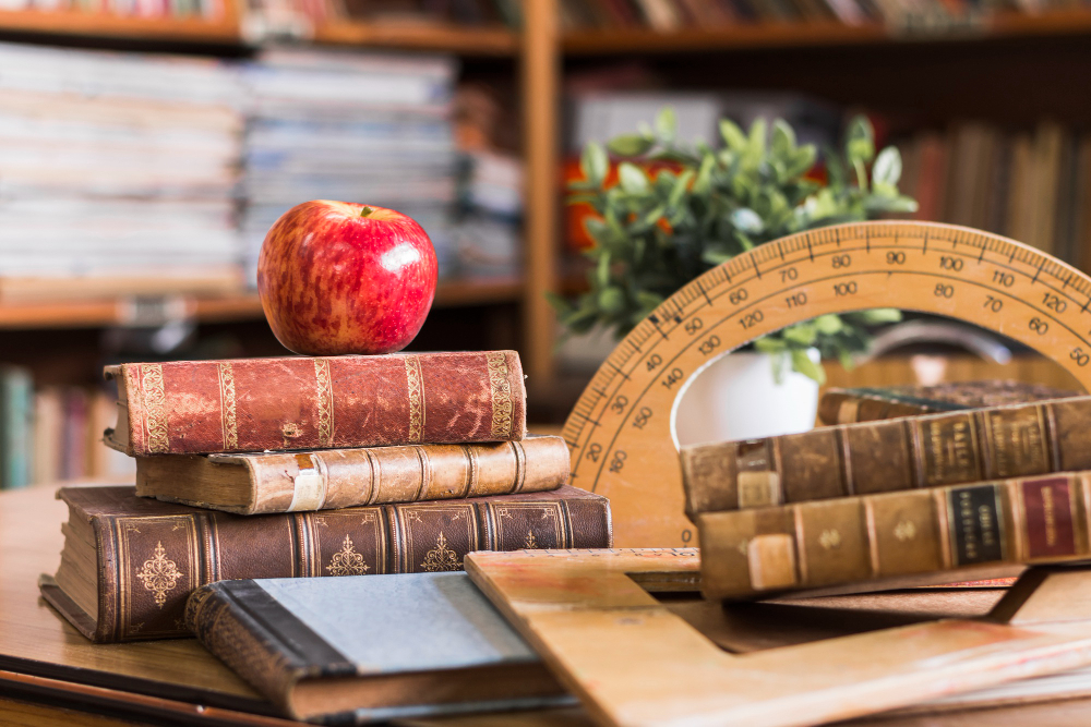 Mesa em uma biblioteca contendo alguns livros antigos empilhados e uma maçã no topo deles, e alguns materiais de ensino também antigos, representando a história da educação e sua evolução ao longo do tempo