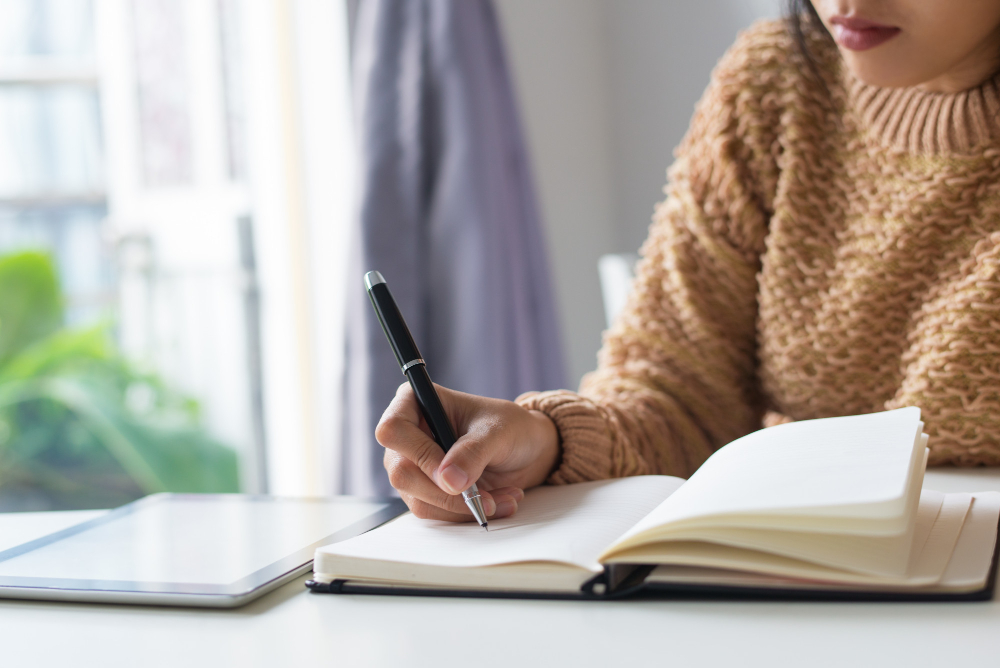 Jovem estudante em sua mesa de estudos, escrevendo em um caderno enquanto se prepara para o ENEM. Ela está estudando tipologia textual para aplicar na redação do exame
