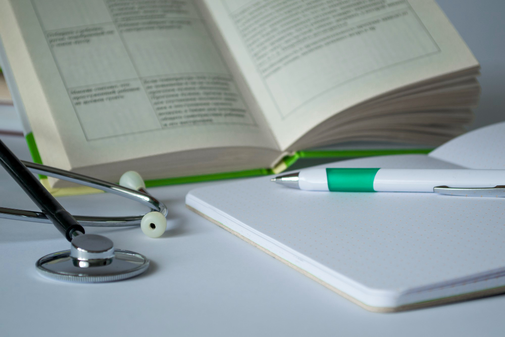 Um livro e um caderno abertos com um estetoscópio ao lado, em cima de uma mesa de estudos. Os objetos representam as disciplinas da Medicina