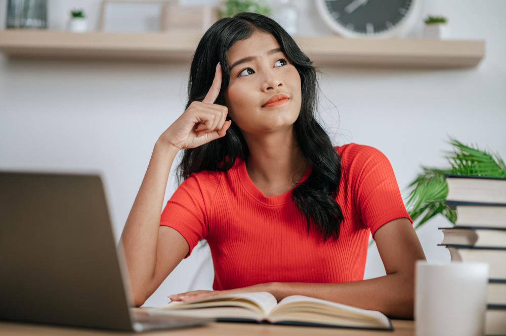 Jovem estudante em sua mesa de estudos, com um notebook e um livro à sua frente. Ela está reflexiva, pensando sobre os erros cometidos no TCC.