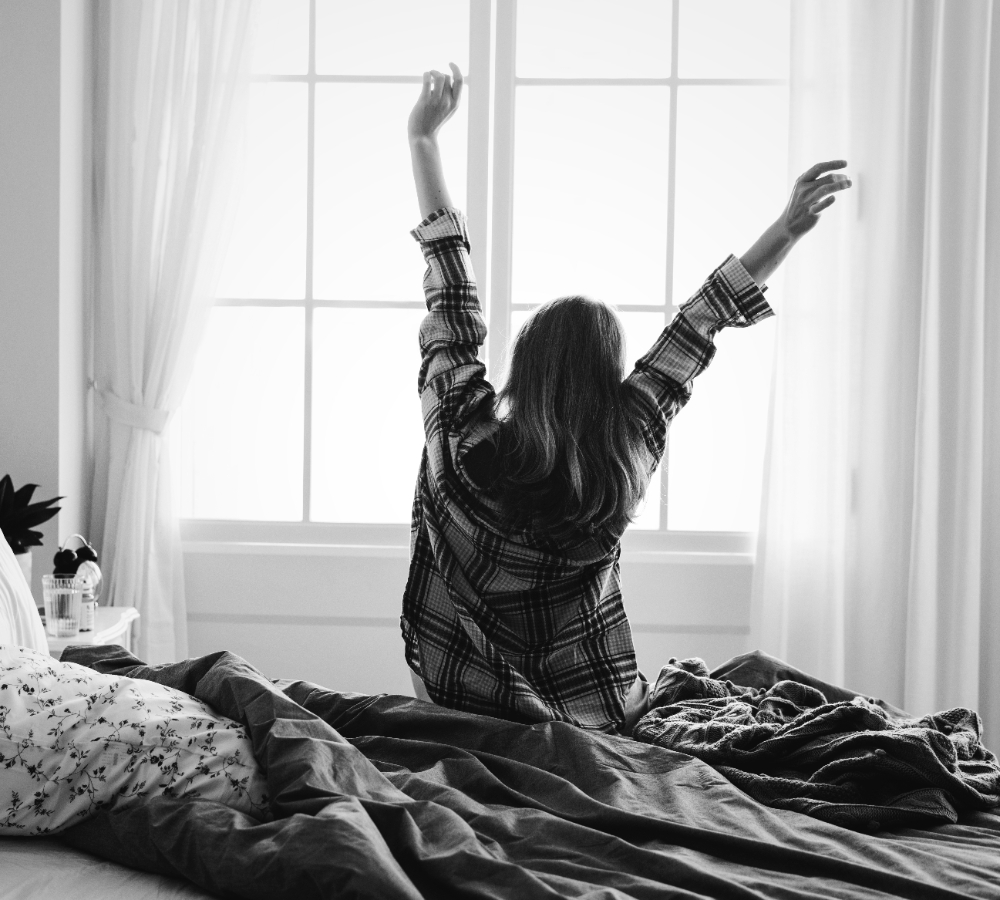 Jovem mulher sentada em sua cama com os braços levantados, espreguiçando-se após acordar cedo