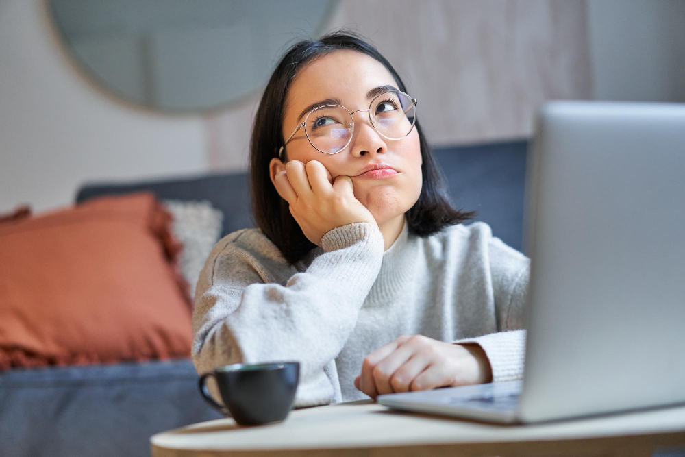Jovem mulher sentada no chão da sala, de frente ao notebook, com uma expressão desanimada durante um momento de tédio.
