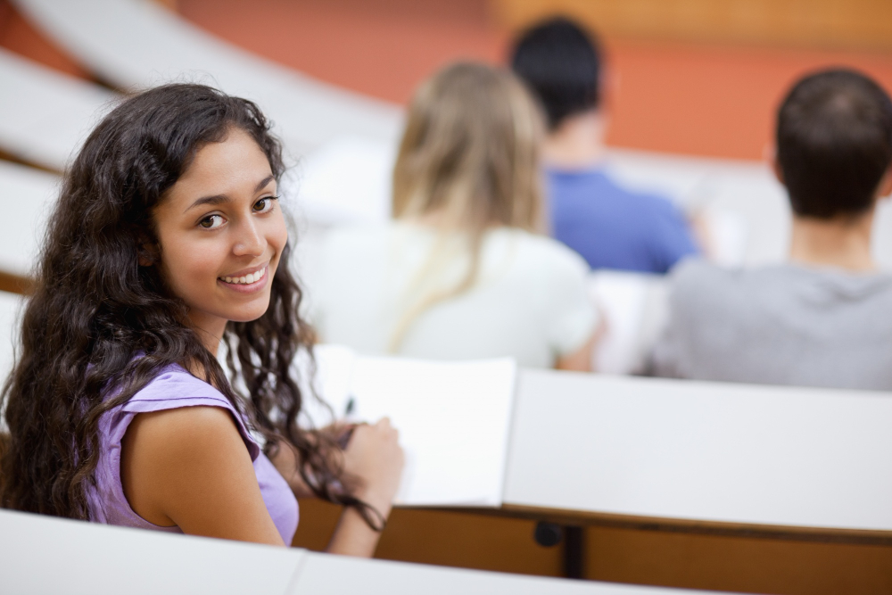 Aluna sentada no banco do auditório de uma faculdade, olhando sorridente para câmera, enquanto estuda para melhorar seu desempenho acadêmico