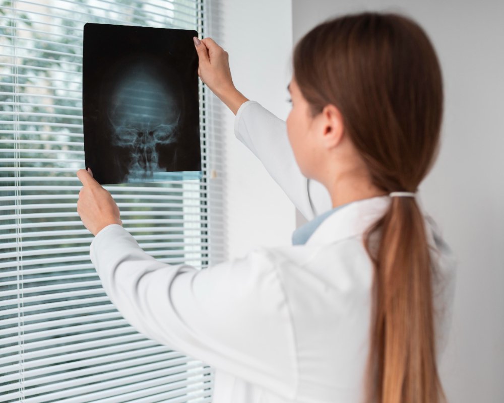 Estudante de medicina em um consultório, de frente para uma janela, analisando um raio-x feito pelo negatoscópio.