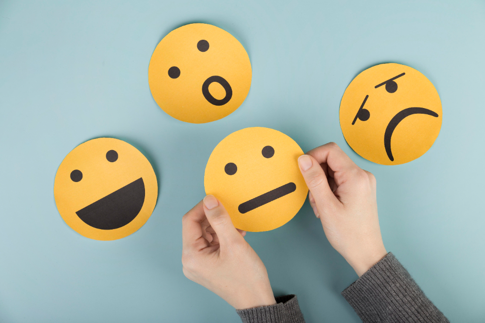 Par de mãos segurando alguns papéis redondos com emojis desenhados, cada um com um sentimente: tristeza, alegria, indiferença e surpresa. Eles representam o que são as emoções