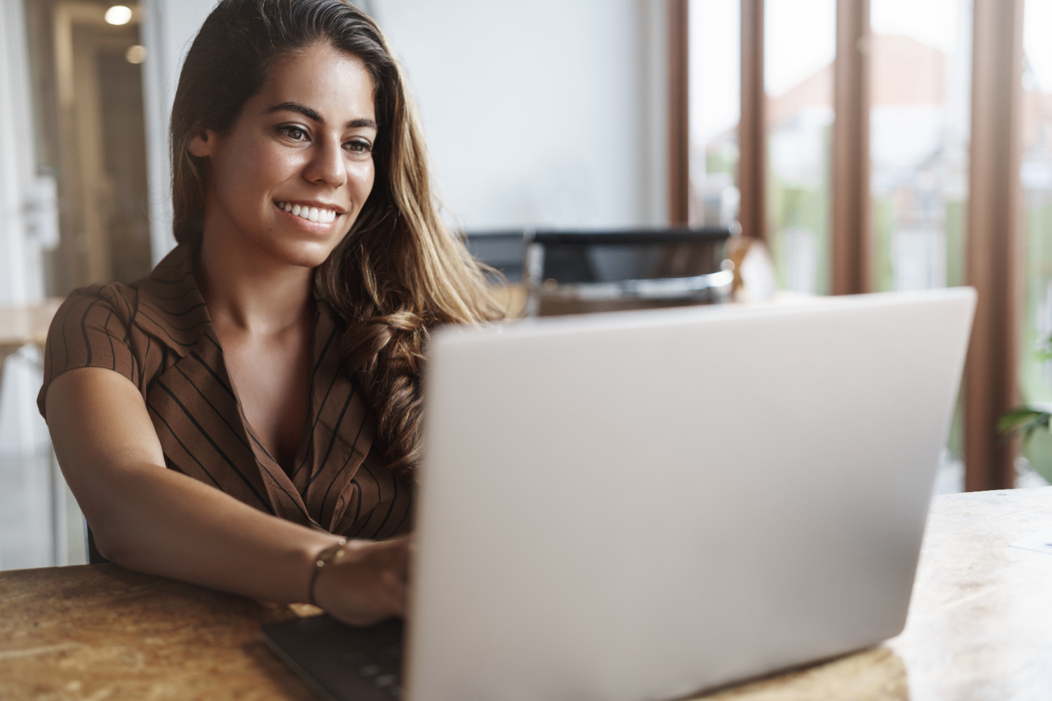 Jovem mulher sorridente, sentada em um dos cômodos de usa casa, utilizando um notebook para trabalhar pela internet