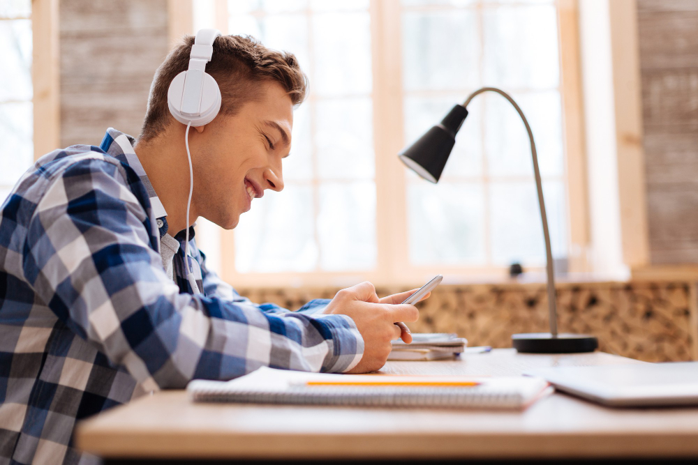 Visão lateral de um jovem sorridente em sua mesa de estudos, utilizando um fone de ouvido enquanto ouve podcasts para estudar