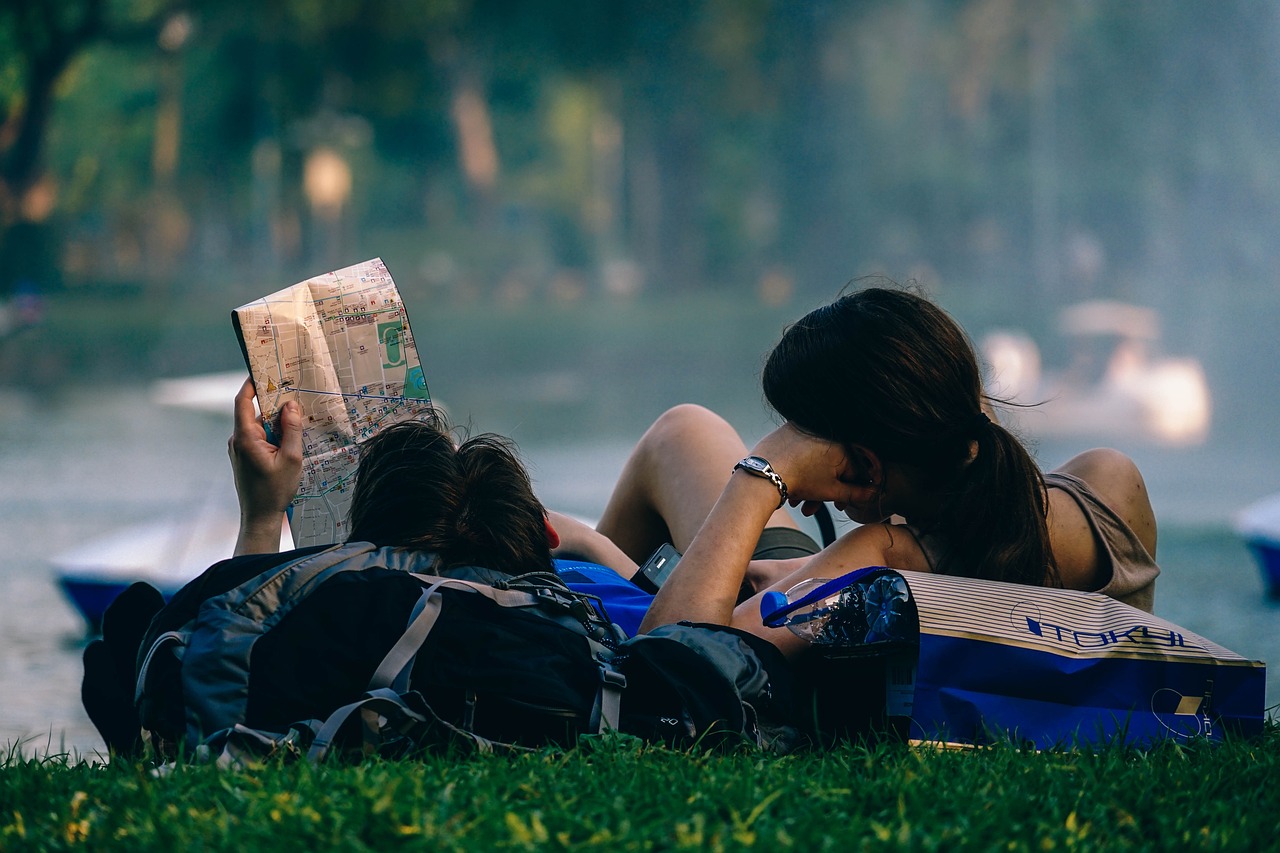 Casal de jovens universitários deitados lado a lado, com as cabeças apoiadas em suas mochilas. O rapaz está segurando um mapa, enquanto eles aproveitam os benefícios das viagens