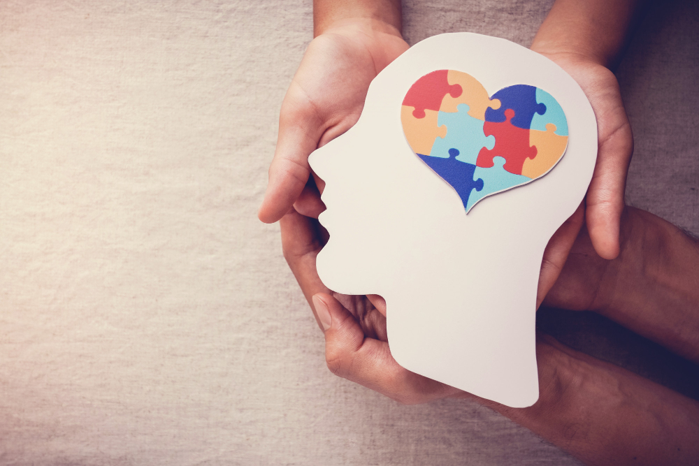 Mãos seguram a silhueta de uma cabeça humana feita de papel, com um coração colorido em formato de quebra-cabeça na região do cérebro, simbolizando o controle emocional e a saúde mental.